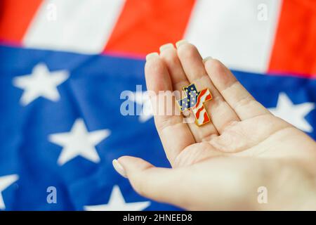 Close up of young woman holding Christian cross pin in her hand. American flag on the background. Patriotism and religious rights concept. Stock Photo