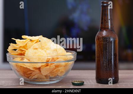 A clear glass bowl full of chips, a bottle of fresh beer and a bottle cap on a wooden table. In the background, out of focus, there is a television wi Stock Photo