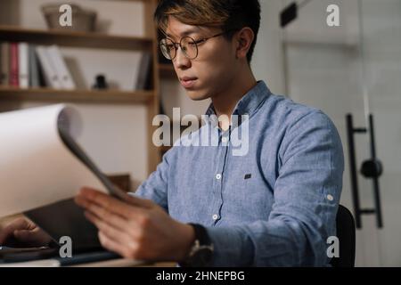 Focused asian guy in eyeglasses working with papers while sitting at table in office Stock Photo