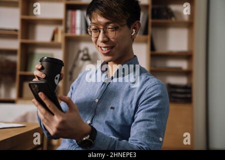 Smiling asian guy in earphones drinking coffee while using mobile phone in office Stock Photo