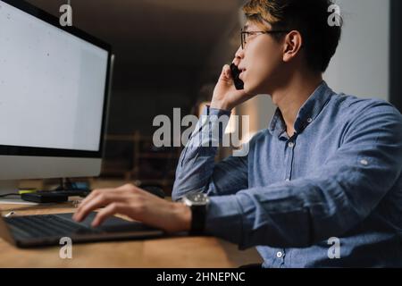 Focused asian guy talking on cellphone while working with computers at office Stock Photo