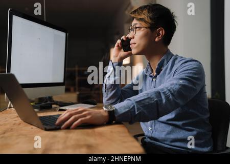 Focused asian guy talking on cellphone while working with computers at office Stock Photo