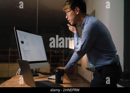 Focused asian guy talking on cellphone while working with computers at office Stock Photo