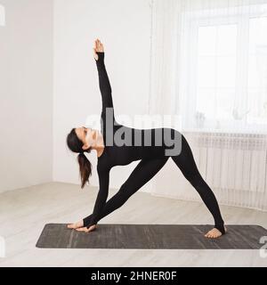 Woman in black sports overalls practicing yoga doing Utthita Trikonasana exercise, extended triangle pose, training while standing on a mat in the roo Stock Photo