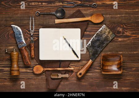 Top view of vintage steak fork, knife and hatchet on wooden table next to notebook. Barbecue appliances and open notepad, food blogging and menu conce Stock Photo