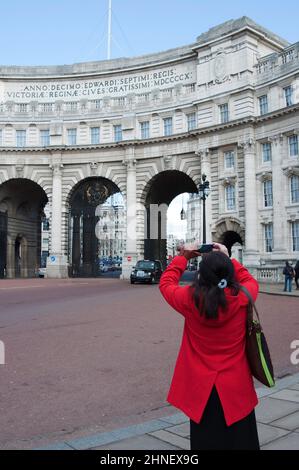 Unrecognizable young woman taking a picture of Admiralty Arch, near Trafalgar Square. Dressed in red, seen from her back. Westminster, London, England Stock Photo