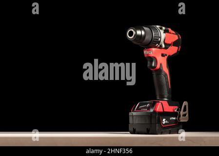 a cordless screwdriver stands on a wooden table on a black background. Cordless drill with lithium-ion battery in red. Professional tool for drilling Stock Photo
