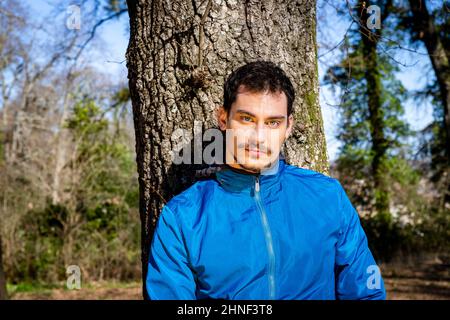 Portrait of a beautiful young man with blue eyes. The man is training in the woods and is leaning against an oak tree. Stock Photo