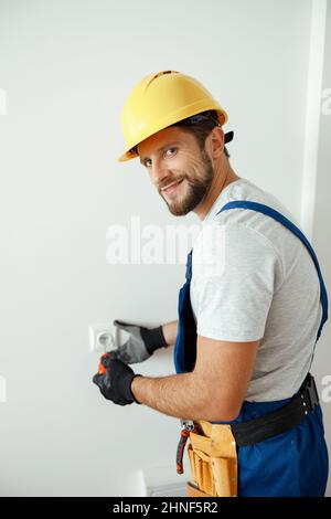 Happy male worker, professional electrician in hard hat and protective gloves smiling at camera while replacing electrical socket in a room Stock Photo