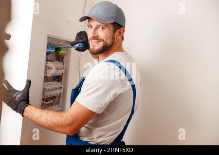 Electrical technician in uniform smiling at camera, using screwdriver while checking fuses in a switchboard on the wall Stock Photo