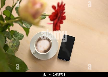 Coffee cup and mobile phone under a flower bouquet on a light wooden table, business concept for constant accessibility, copy space, top view from abo Stock Photo