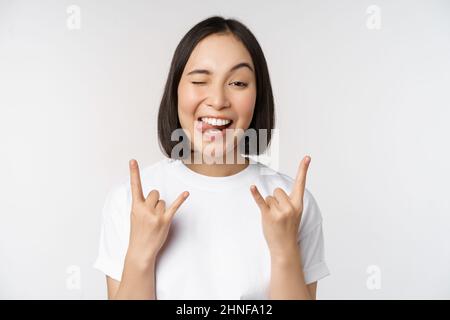 Sassy asian girl shouting, enjoying concert or festival, showing rock on, heavy metal sign, having fun, standing over white background Stock Photo