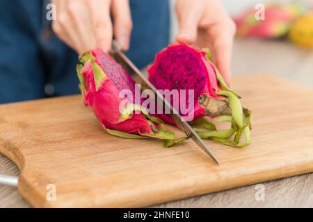 Female hands is cutting a dragon fruit or pitaya with pink skin and pulp with black seeds on wooden cut board on the table. Exotic fruits, healthy Stock Photo
