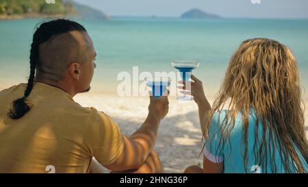 The love couple is holding a glass of blue curacao cocktail, on sea. Man with plait and blonde woman drink alcohol on sand beach in shadow by azure ocean of a tropical island. Summer holiday concept. Stock Photo