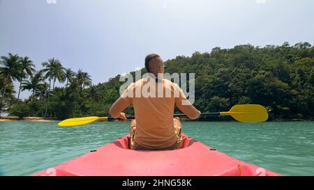 Young man with sunglasses and hat rows pink plastic canoe along sea against green hilly islands with wild jungles. Traveling to tropical countries. Strong guy is sailing on kayak in ocean, back view. Stock Photo