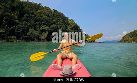 Young man with sunglasses and hat rows pink plastic canoe along sea against green hilly islands with wild jungles. Traveling to tropical countries. Strong guy is sailing on kayak in ocean, front view. Stock Photo