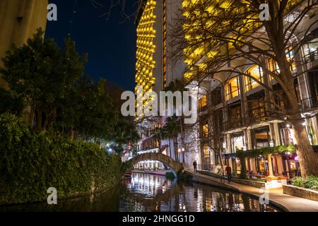 The popular riverwalk in a night scene in downtown San Antonio, Texas, with an arch bridge and tall buildings along the river reflected in the water. Stock Photo