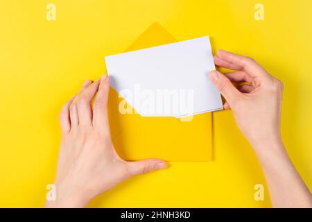 Top view on Hands put the card into the envelope. Blank white card with yellow envelope isolated on yellow background. Template or mock-up. Banner Stock Photo