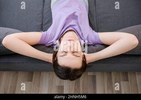 Top view view of peaceful calm young woman relaxing on cozy grey couch, enjoying shiny morning alone at home. Mindful millennial lady resting on comfo Stock Photo