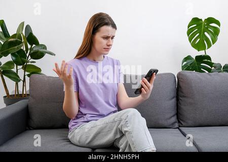 Stressed millennial lady thinking over sudden problems or relations break up after reading sms on cellphone. Upset young woman 20s sitting on couch Stock Photo