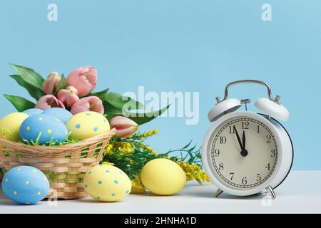 Easter concept. Basket of eggs alarm clock flowers on a blue background copy space. Stock Photo
