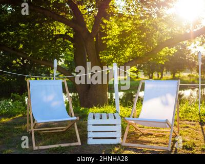 two white beach seat chairs and wooden box as a table between it on nature on a lake shore, city break and place for relax Stock Photo