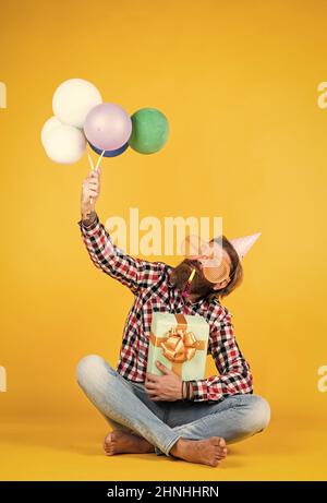 Caucasian mature hipster with funny party glasses in checkered shirt holding balloons and gift box, celebration Stock Photo