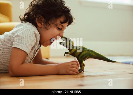 Adventurous young boy imitating a dinosaur toy while lying on the floor in his play area. Creative little boy having fun during playtime at home. Stock Photo