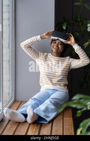 Happy smiling afro female sitting on windowsill at home, enjoying VR experience Stock Photo