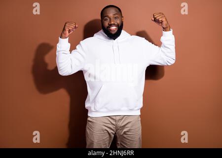 Photo of sweet strong guy wear white hoodie smiling rising fists showing power isolated brown color background Stock Photo
