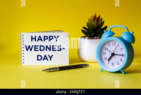 Notepad with text Happy Wednesday on yellow background with clock. Stock Photo