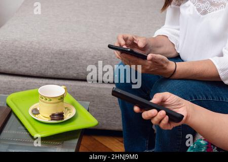 close-up of caucasian female hands in an apartment living room holding a telephone and a television control, with a cup of coffee and a chocolate alfa Stock Photo