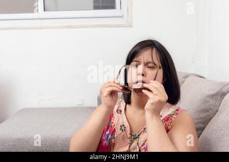 adult woman with down syndrome latin caucasian sitting on the sofa in the living room of her apartment putting on sunglasses, fashion concept with cop Stock Photo
