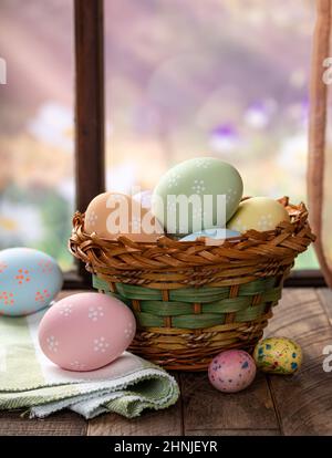 Colorful easter eggs in a basket and on a rustic wooden table by a window with rural background Stock Photo