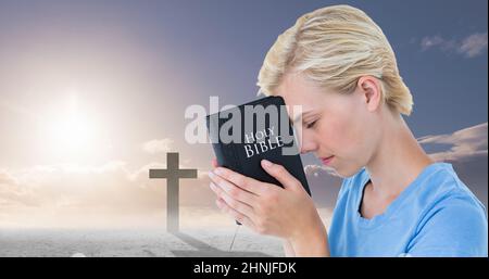 Close-up of caucasian young woman with bible praying and silhouette cross against sky Stock Photo