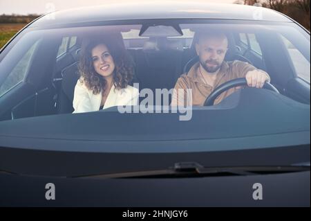 Front view in black car of focused male driver and smiling female passenger traveling by car out the town. Stock Photo