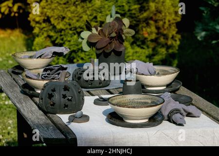 Rustic table setting outside in garden with empty craft ceramic tableware, black plates and rough bowls, pumpkin decorations, on linen tablecloth over Stock Photo
