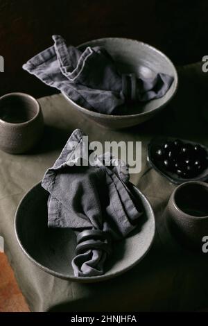 Rustic table setting with empty craft ceramic tableware, grey rough bowls and cups on linen tablecloth. Dark background Stock Photo
