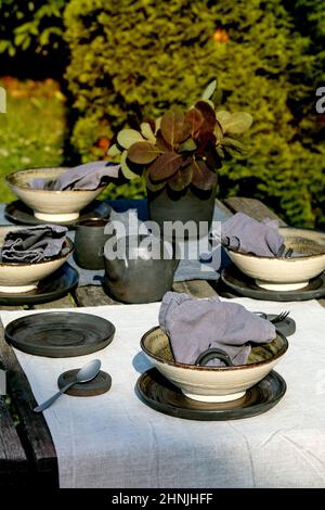 Rustic table setting outside in garden with empty craft ceramic tableware, black plates and rough bowls, teapot on linen tablecloth over old wooden ta Stock Photo