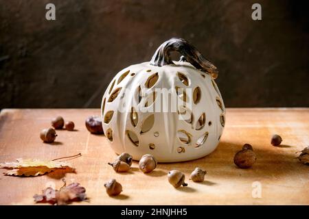 Halloween or Thanksgiving decorations, white handcrafted carved ceramic pumpkin standing on orange stone table with autumn leaves and acorns. Hallowee Stock Photo