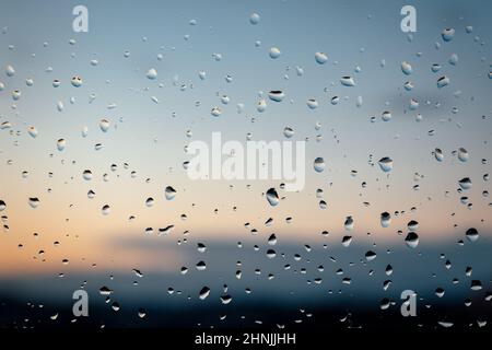 Raindrops after a storm on window at sunset. The raindrops are in focus and the background is blurred. The sunset has orange and pink hues. Stock Photo