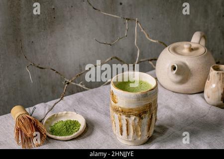 Traditional japanese hot green frothy tea matcha in handmade ceramic cup, powdered matcha, teapot and bamboo whisk standing on grey linen tablecloth. Stock Photo