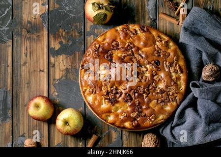 Homemade apple and walnuts cake Charlotte on plate with fresh gardening apples, cinnamons and nuts around over dark wooden plank background. Autumnal Stock Photo