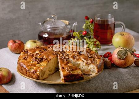 Homemade apple and walnuts cake Charlotte on plate with fresh gardening apples, nuts, rose hip tea around on grey linen table cloth. Autumnal home bak Stock Photo