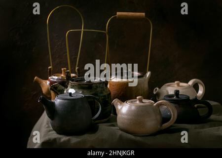Variety of craft handmade ceramic teapots kettles for tea ceremony standing on linen tablecloth in dark room. Stock Photo