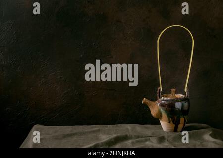 Craft handmade ceramic teapot kettle for tea ceremony standing on linen tablecloth in dark room. Stock Photo