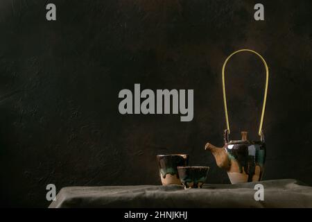 Craft handmade ceramic teapot kettle with cup of hot green tea for tea ceremony standing on linen tablecloth in dark room. Stock Photo