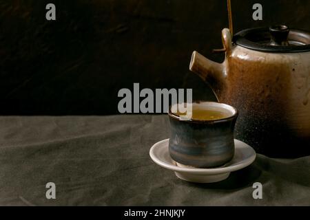Craft handmade ceramic teapot kettle with cup of hot green tea with saucer, standing on linen tablecloth in dark room. Close up Stock Photo