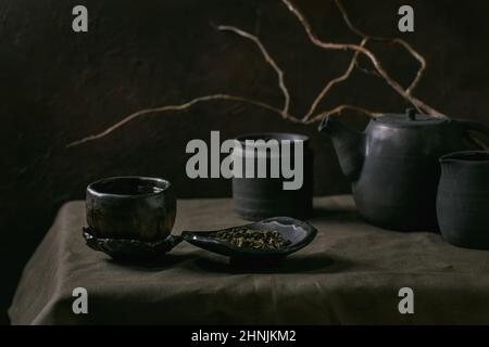 Set of craft handmade black ceramic teapot kettle, jug, jar with tea leaves, wabi sabi cup, saucer for tea ceremony standing on linen tablecloth with Stock Photo