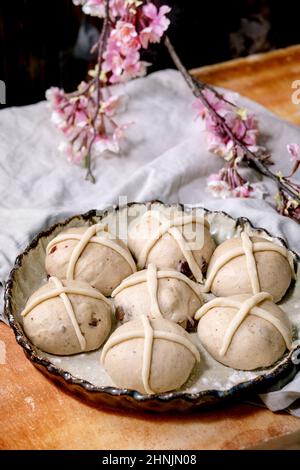 Raw unbaked buns. Ready to bake homemade Easter traditional hot cross buns in ceramic baking dish over orange stone table with linen cloth and pink fl Stock Photo